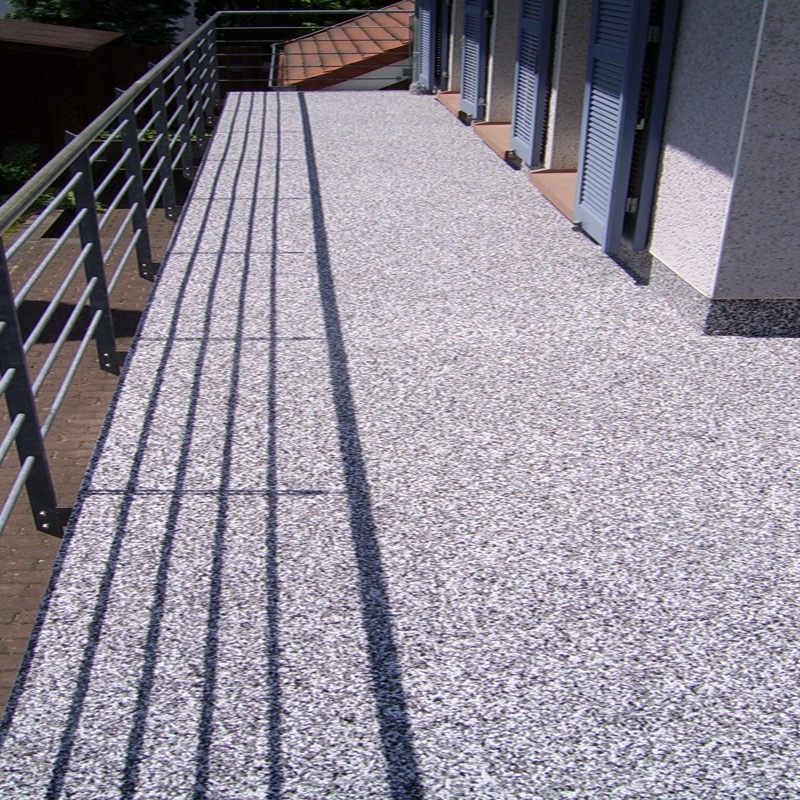 Ein Balkon oder eine Terrasse mit einem grobkörnigen Bodenbelag. Die Schatten von einem Geländer fallen auf diesen Boden. An der rechten Seite sind Fenster oder Türen mit Jalousien sichtbar.